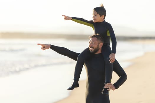 Little son on the shoulders of his father pointing to the sea. Mid shot