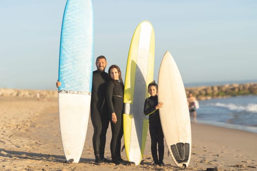A family of surfers standing on the seashore in wetsuits. Mid shot