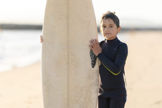 A little boy in a wetsuit holding a surfboard on the seashore. Mid shot