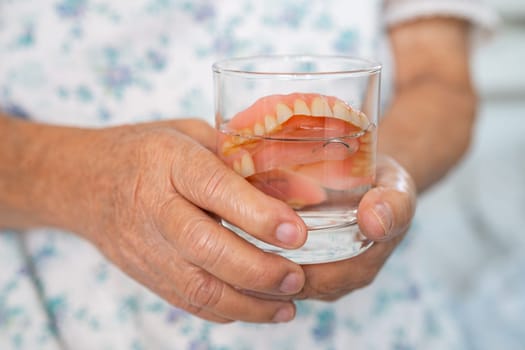 Asian senior woman patient holding and washing denture in water cleanser glass for good chewing.