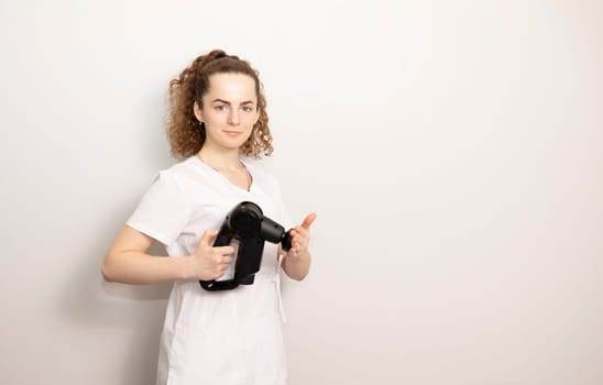 Rehabilitation Specialist, Physical Therapist In White Medical Clothes Holds Massage Gun, Massager Product, Copy Space For Text. Health Specialist, Rehabilitation. Horizontal plane.