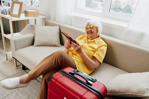 Happy senior woman with passport and travel ticket packed a red suitcase, vacation and health care. Smiling old woman joyfully sitting on the sofa before the trip raised her hands up in joy. High quality photo