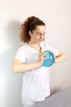 Caucasian Rehabilitation Specialist, Physiotherapist In White Medical Clothes Holds Massage Ball With Pimples In Therapeutic Cabinet. Health Specialist, Rehabilitation. Vertical. High quality photo