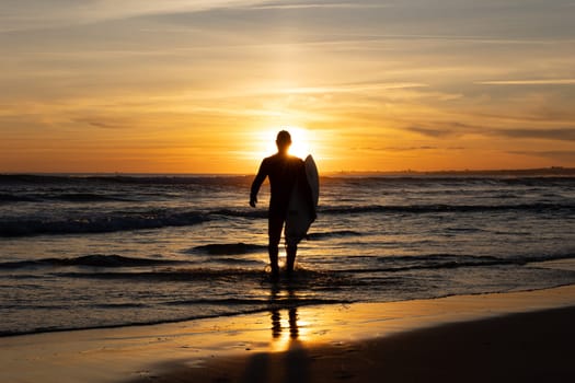 Dark silhouette of a man surfer walking out on the seashore holding a surfboard at bright sunset. Mid shot