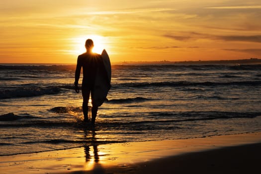 Dark silhouette of a man surfer holding surfboard at bright sunset. Mid shot