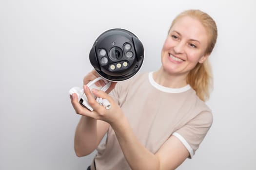 Smiling Caucasian Woman Holds IP Smart Outdoor Security Camera On White Grey Background. Smart Home, House Surveillance, System With Optical Zoom, Water Proof Cover, Facial Recognition. Horizontal.