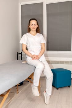 Smiling Young Rehabilitation Specialist, Physician In White Medical Clothes Sitting On Couch In Therapeutic Cabinet. Health Specialist, Rehabilitation. Vertical Plane. High quality photo