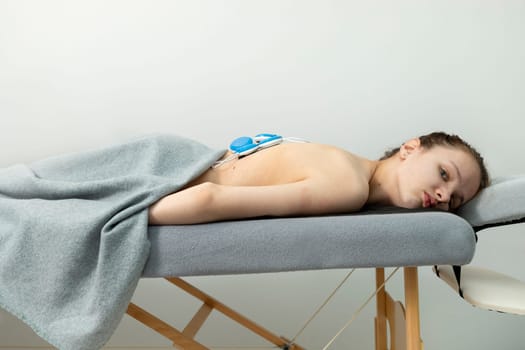 Caucasian Girl With Cerebral Palsy, Scoliosis Lying on Couch, Getting Wireless Electric Muscle Stimulator Massage. Pain Treatment of Woman's Back. Rehabilitation, Medical Equipment. Horizontal plane.