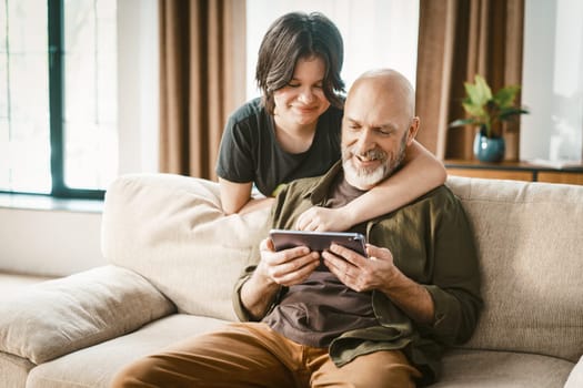 Father and daughter spend quality time together playing online games. Senior man sits comfortably on sofa while teenager affectionately hugs him, showcasing their strong family connection. . High quality photo