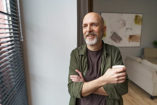 Happy and smiling mature senior man standing near window, holding cup of coffee, and gazing outside. Moment of contentment and relaxation in senior man's life. High quality photo