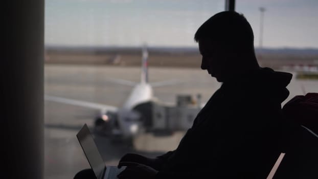 A young man in a sweatshirt is sitting at the airport with a laptop. An IT specialist works sitting in an airport. Silhouette of a man on the background of the planes outside the window. 4k