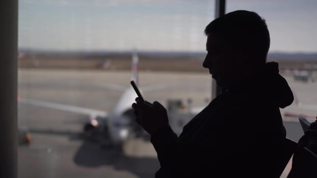 A young man in a sweatshirt is sitting at the airport with a phone in his hands. The man is using a smartphone. Silhouette of a man on the background of the planes outside the window. 4k