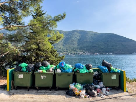 Overflowing garbage cans stand in a fence on the seashore near a tree. High quality photo