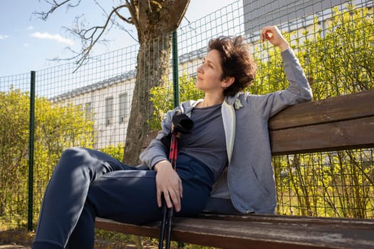 Pretty Woman Sits on Bench Holding Sticks After Nordic Walking. Beautiful Happy Caucasian Woman Enjoys Sunny Day. Physical Activity With Walking Poles. Sports Tourism, Outdoor Recreation. Horizontal.