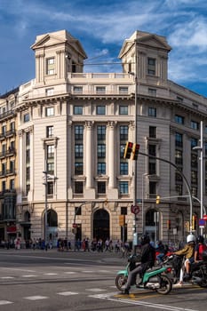 Crossroads in Barcelona with view of a building, in the evening, sunset. Motorcyclists are waiting at the traffic lights, people are crossing the road.