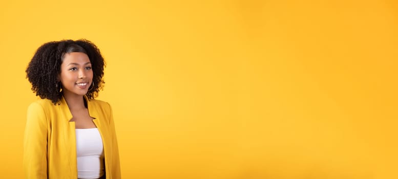 Pretty young african american curly woman looking at empty space and smiling, posing isolated on yellow studio background. Huge sale, good news, fashion lifestyle and beauty care