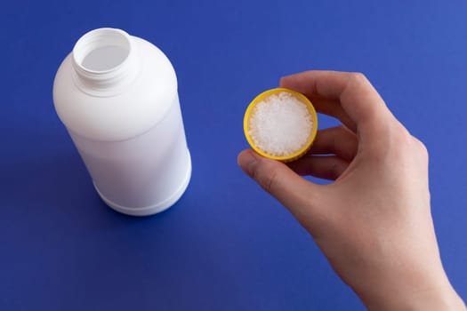 Woman Hand Holds Dosage Of Pipe Cleaner Granules. Removal Of Blockage In Sink, Shower With Special Remedy With Chemicals. Cleaning Clog In Kitchen And Bathroom. White Plastic Bottle On Background.