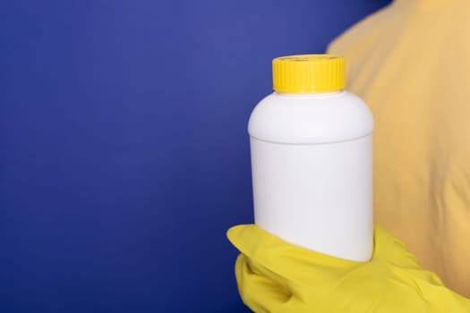 Woman Hand In Yellow Gloves Holds White Bottle With Pipe Cleaner Granules. Removal Of Blockage In Sink, Shower With Special Chemicals. Cleaning Clog In Kitchen, Bathroom. Blue On Background,Copyspace