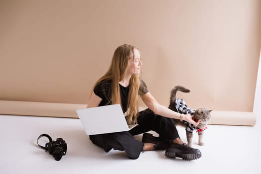 A business woman freelancer in a black is working, typing on a laptop apple and sitting on a white floor. blonde assistant in video production with computer MacBook and camera with cat