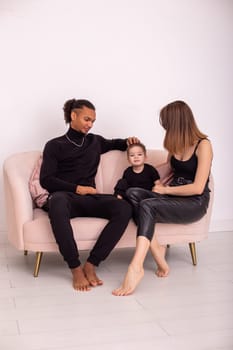 Beautiful multiracial family with little boy, in black casual clothes, sits in the light living room on light pink sofa, communicate. Vertical. Copy space