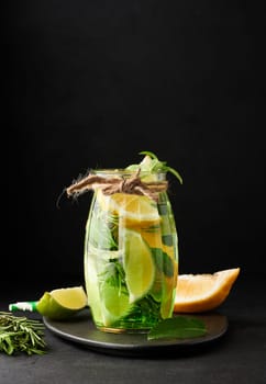 Lemonade in a transparent glass with lemon, lime, rosemary sprigs and mint leaves on a black background