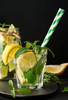 Lemonade in a transparent glass with lemon, lime, rosemary sprigs and mint leaves on a black background