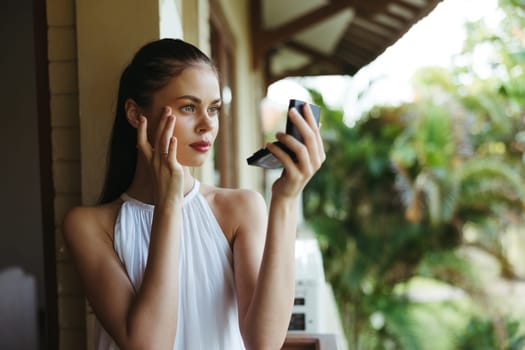 A young beautiful woman looks at her face with makeup in the mirror outside in the summer, protective spf products for problem skin with acne. High quality photo