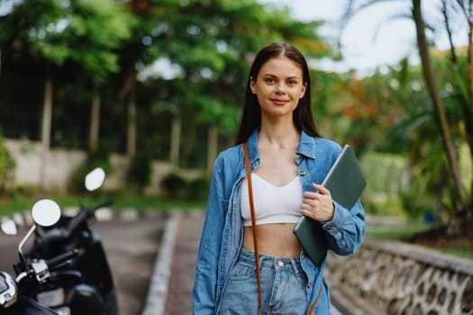 Woman smiling walking in the park outside with laptop freelancer against a backdrop of green palm trees in summer, tropical backdrop, blogger on a trip, work online. High quality photo