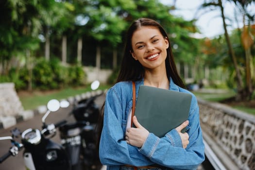 Woman smiling walking in the park outside with laptop freelancer against a backdrop of green palm trees in summer, tropical backdrop, blogger on a trip, work online. High quality photo