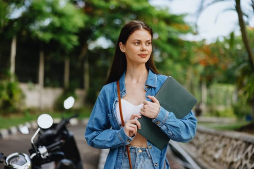 Woman smiling walking in the park outside with laptop freelancer against a backdrop of green palm trees in summer, tropical backdrop, blogger on a trip, work online. High quality photo