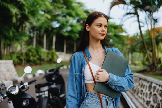 Woman smiling walking in the park outside with laptop freelancer against a backdrop of green palm trees in summer, tropical backdrop, blogger on a trip, work online. High quality photo