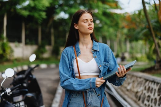 Woman smiling walking in the park outside with laptop freelancer against a backdrop of green palm trees in summer, tropical backdrop, blogger on a trip, work online. High quality photo