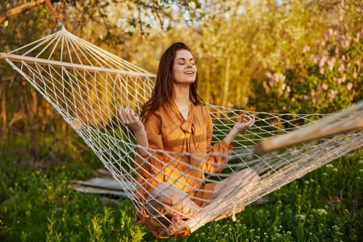 a happy woman in a long orange dress is resting lying in a hammock at the dacha, meditating with her eyes closed, illuminated by the summer sun during sunset. High quality photo