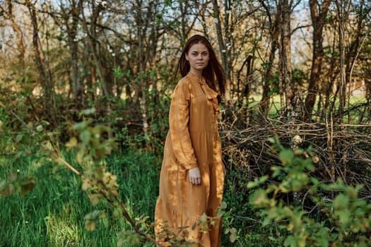 a woman with long hair walks in the shade near the trees, dressed in a long orange dress, enjoying the weather and the weekend, while looking at the camera. The theme of privacy with nature. High quality photo