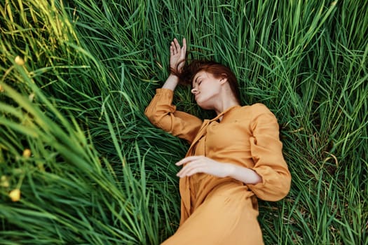 a close horizontal photo of a pleasant woman in a long orange dress resting lying in the tall grass with her eyes closed in sunny weather. High quality photo