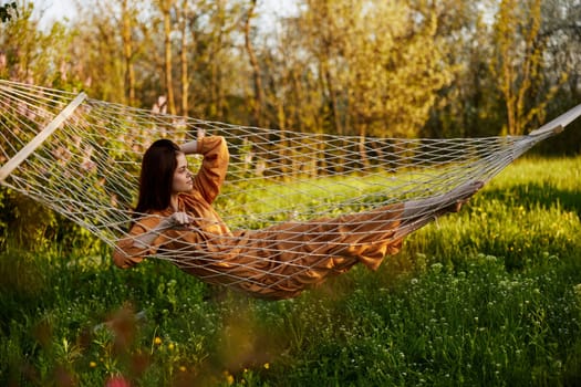an elegant, slender woman is resting in nature lying in a mesh hammock in a long orange dress enjoying the rays of the setting sun on a warm summer day looking away. High quality photo