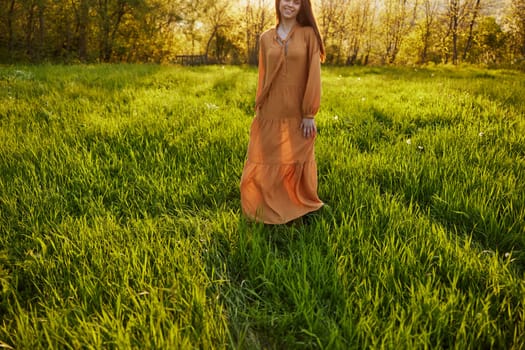 horizontal photo of a woman in an orange dress photographed without a face, standing in a green field during sunset, illuminated from the back. High quality photo