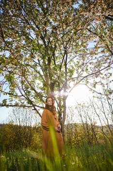 a sweet, modest woman with long hair stands in the countryside near a flowering tree and looks at the camera with her hands clasped together. High quality photo