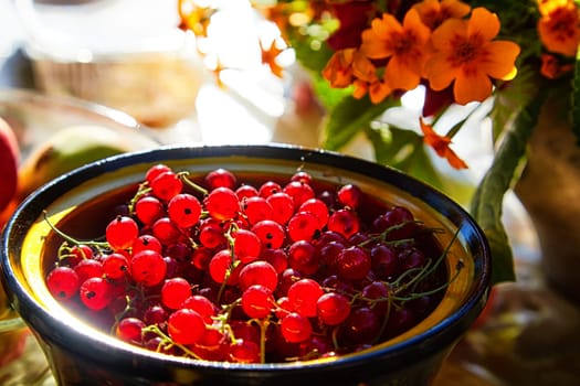 Red currant berries in a wooden plate on the table and a blurred background with red flowers