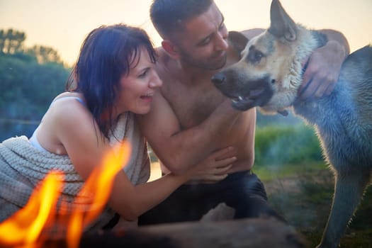 Happy wet couple relaxin, having fun and hugs with big dog near fire in camping on nature in summer sunny evening in sunset. Family or lovers have date and rest outdoor. Concept of love