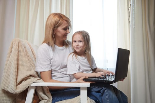 Beautiful young woman and her little cute daughter are using laptop at home. Enjoying spending time together with Internet and modern technologies