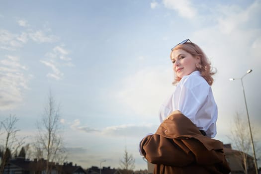 Beautiful girl with red hair in short skirt, white shirt and raincoat in village or small town. Tall young slender woman and houses and sky with clouds on background on autumn, spring or summer day