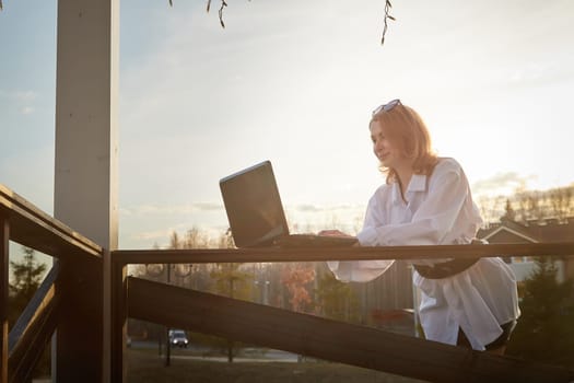 Beautiful redhead moden girl working by laptop in gazebo on autumn, summer or spring day. Businesswoman, student, freelancer or manager works outdoors in natural landscape in a village, city or town