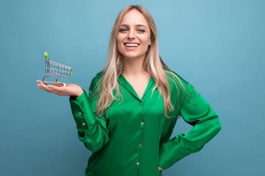 stylish european woman shopper in green shirt with supermarket trolley on blue studio background.