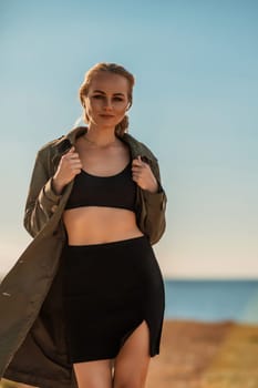 Portrait blonde sea cape. A calm young blonde in an unbuttoned khaki raincoat walks along the seashore, under a raincoat a black skirt and top.