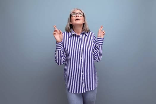 60s middle-aged woman with gray hair in a trendy shirt crossed her fingers on a bright studio background with copyspace.