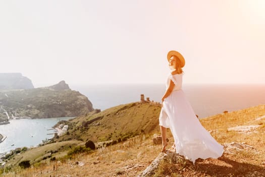 Happy woman in a white dress and hat stands on a rocky cliff above the sea, with the beautiful silhouette of hills in thick fog in the background