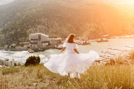 Happy woman in a white dress and hat stands on a rocky cliff above the sea, with the beautiful silhouette of hills in thick fog in the background