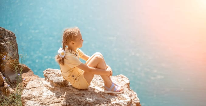 Happy girl perched atop a high rock above the sea, wearing a yellow jumpsuit and braided hair, signifying the concept of summer vacation at the beach