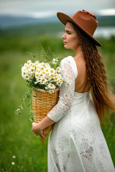 A middle-aged woman in a white dress and brown hat stands on a green field and holds a basket in her hands with a large bouquet of daisies. In the background there are mountains and a lake